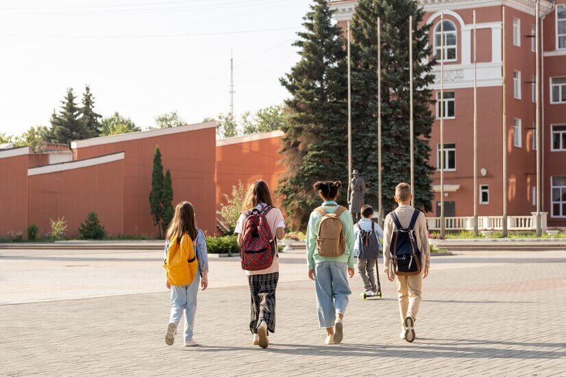 Five children walk together in a school courtyard, each carrying backpacks, surrounded by greenery and buildings in the background.