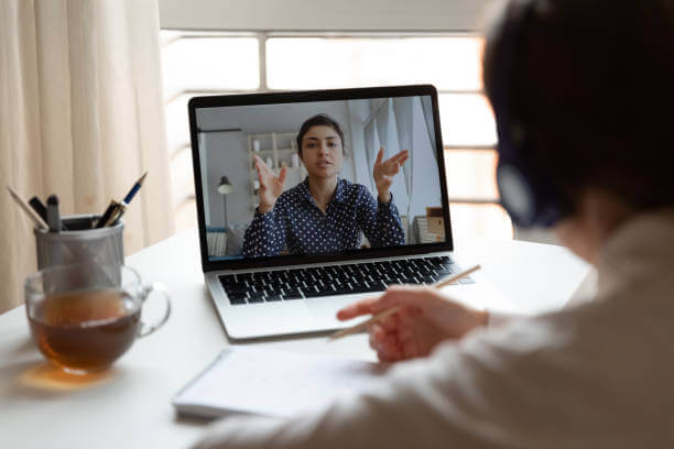 Person participating in a video call on a laptop, engaged in discussion, with a cup of tea and notepad on the table.
