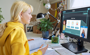 A person in a yellow hoodie is focused on a computer screen displaying an educational interface, with a plant and study materials around.
