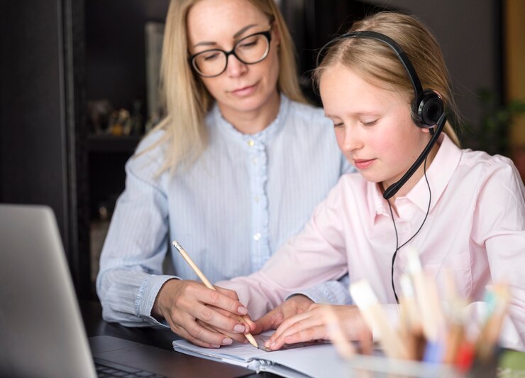 A child wearing headphones is engaged in a learning activity with an adult, both focused on a notebook and a laptop.
