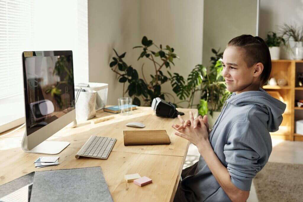 A person sits at a wooden desk with a computer, surrounded by green plants and stationery, in a bright, modern workspace.