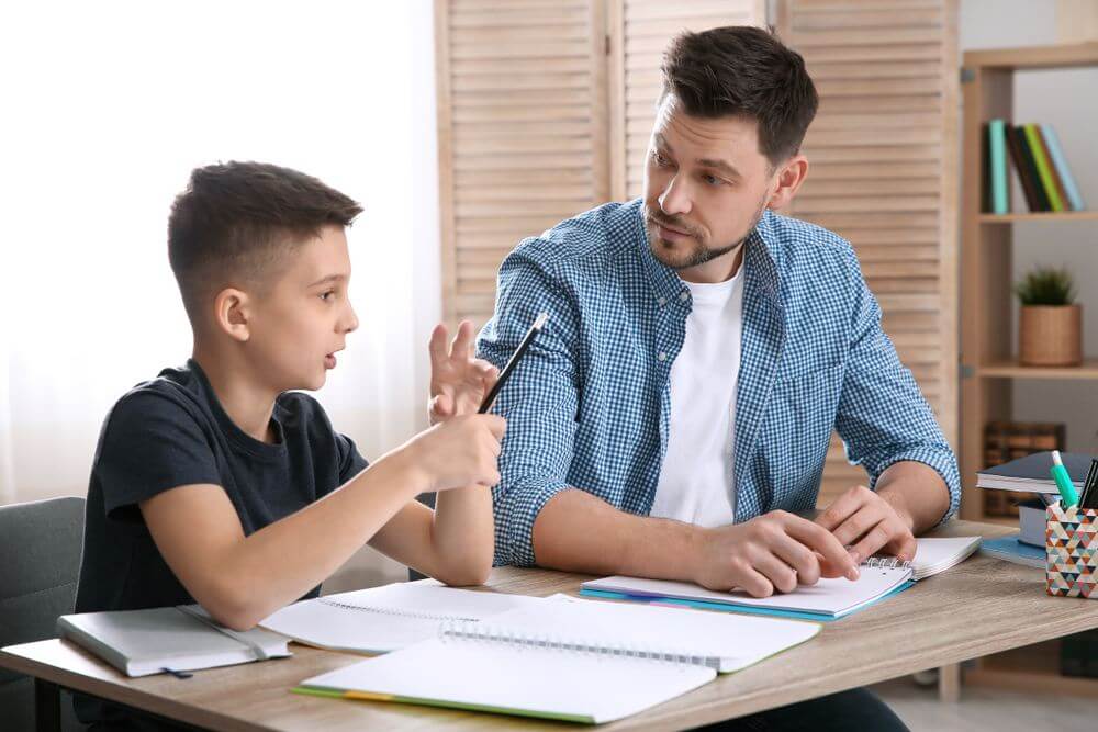 A young boy and an adult sit together at a desk covered with notebooks and stationery, engaged in conversation. Natural light filters through a window.