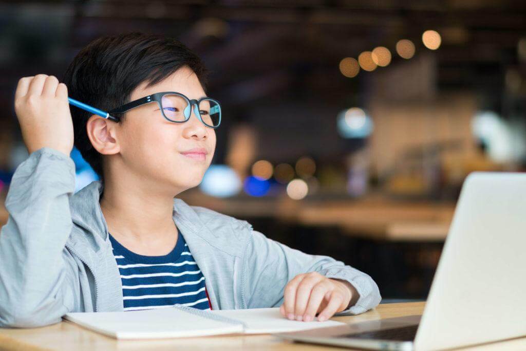 A young person sits at a desk with a laptop, holding a pen and looking thoughtfully at a notebook in a cozy, modern café.