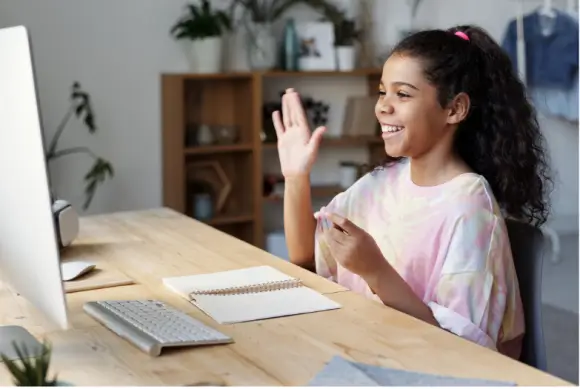 A girl sits at a desk with a computer, gesturing with her right hand, while a notebook and a keyboard are on the table.