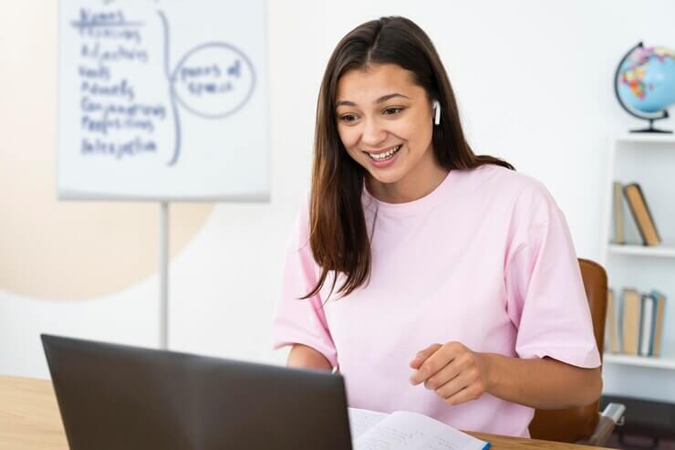 A person wearing pink sits at a desk with a laptop, engaged in online learning, with a whiteboard and a globe in the background.