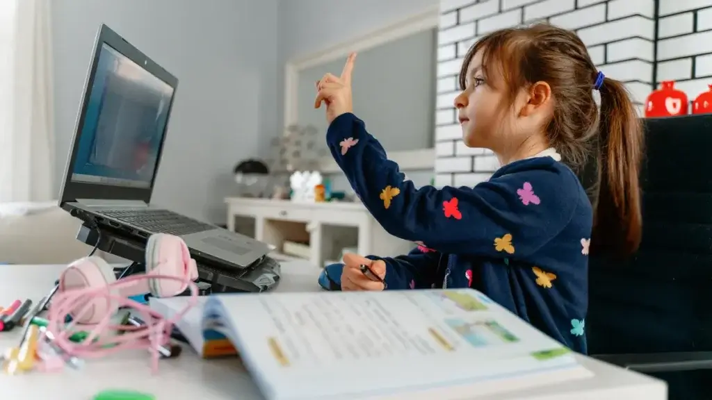 A child raises her hand while sitting at a desk with a laptop, surrounded by school supplies and an open textbook.