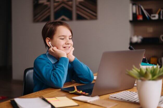 A young child wearing earbuds sits at a wooden desk, focused on a laptop, surrounded by stationery and a potted plant.