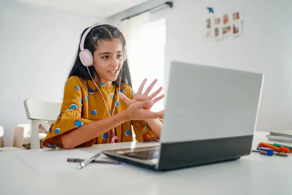 A child in a polka dot shirt, wearing pink headphones, gestures while engaged in a virtual class on a laptop at a bright table.