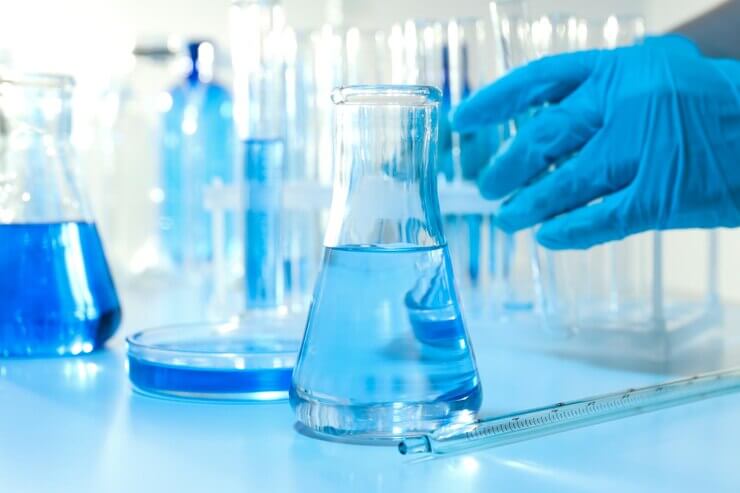 A laboratory scene featuring a blue liquid-filled flask, a petri dish, and a hand in a blue glove reaching for lab equipment.