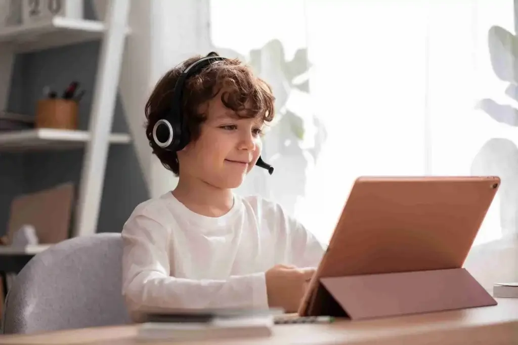A child wearing headphones sits at a desk with a tablet, engaged in an online activity in a bright, modern workspace.