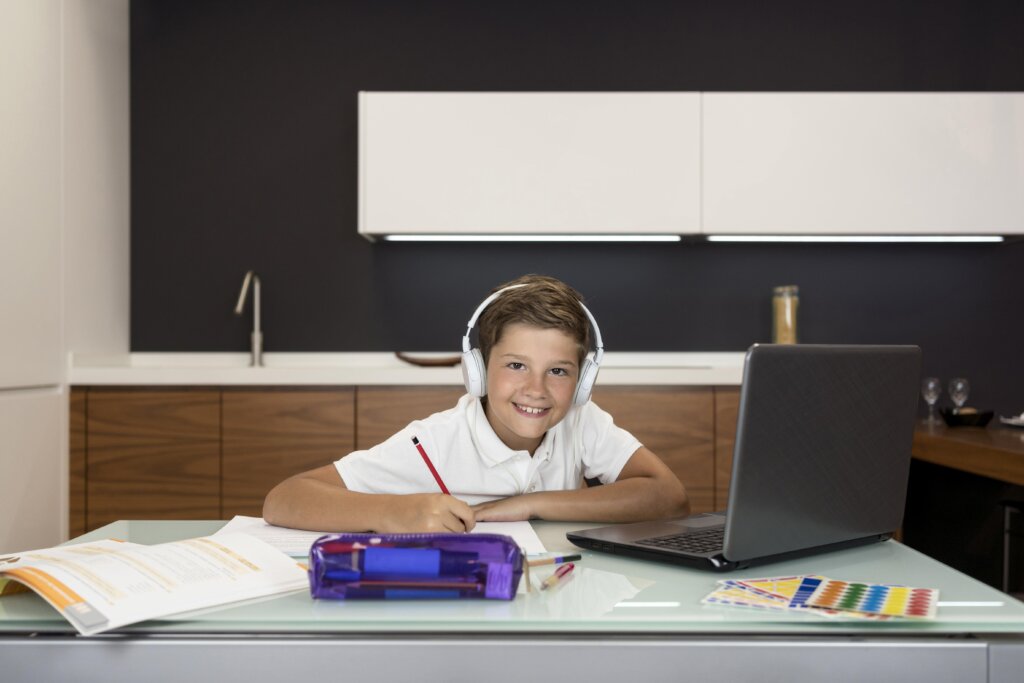 A child wearing headphones sits at a table, focused on homework with a laptop and stationery scattered around.