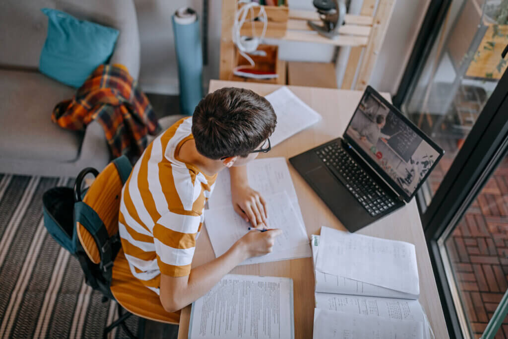 A student in a striped shirt writes notes at a desk with a laptop, surrounded by printed materials and a cozy living space.