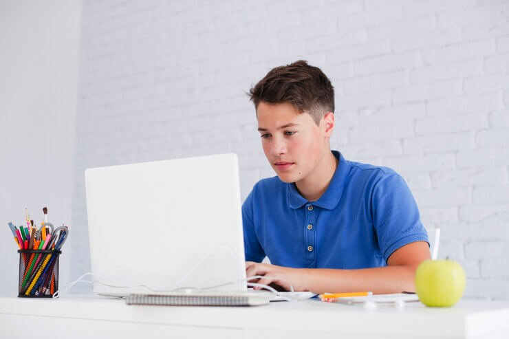 A young man in a blue polo shirt sits at a white desk, focused on a laptop, with a green apple and stationery nearby.