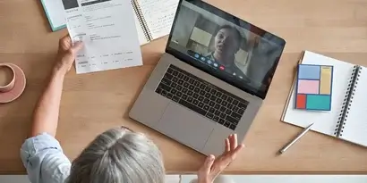 An older person on a video call with a young professional, reviewing documents and notes at a wooden desk.