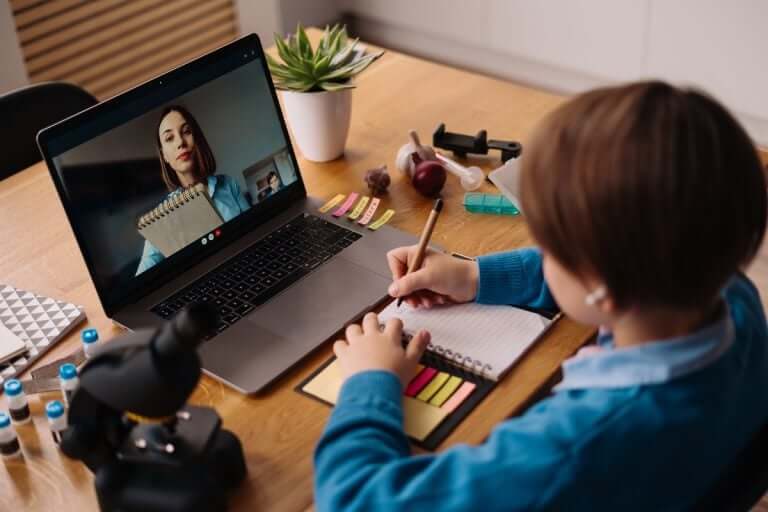 A student sits at a wooden desk, taking notes with a pencil during a video call, surrounded by study materials and a microscope.