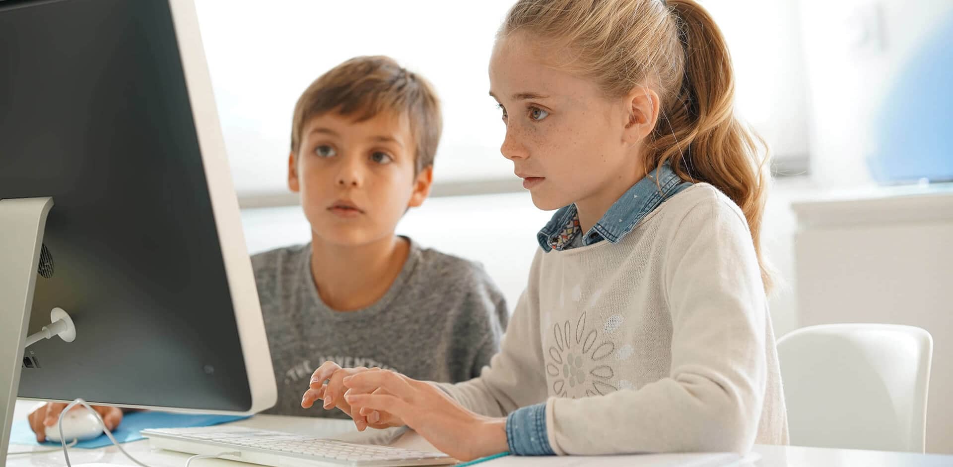 Two children are focused on a computer screen, with one typing on the keyboard while the other observes attentively.