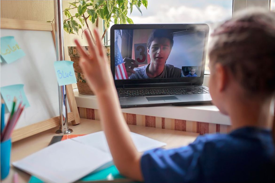 A girl waves at a laptop screen during a video call, surrounded by colorful notes and a plant on a desk by a window.