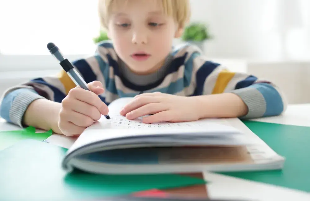 A child wearing a striped sweater writes with a pen on a notebook filled with letters and numbers, surrounded by colorful paper.
