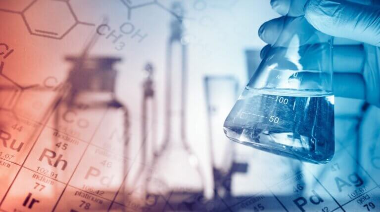 A gloved hand holds a flask filled with a clear liquid, with laboratory equipment and a periodic table softly blurred in the background.