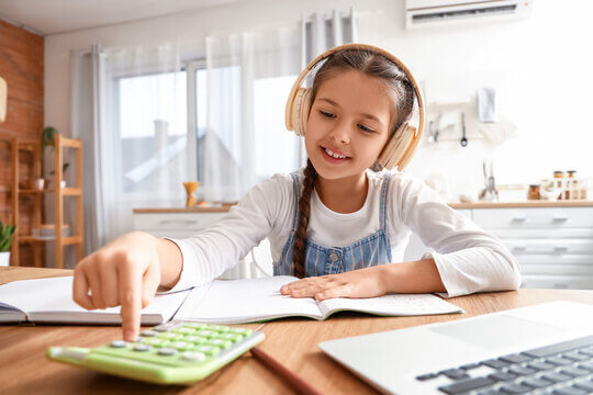A child wearing headphones studies at a wooden table, focused on a calculator, with open notebooks and a laptop nearby.