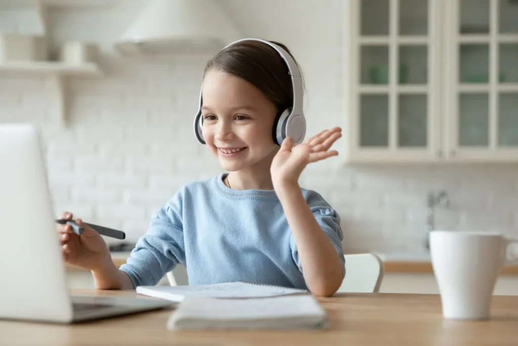 A child in a blue sweater wears headphones while gesturing with one hand, seated at a desk with a laptop and notebooks.