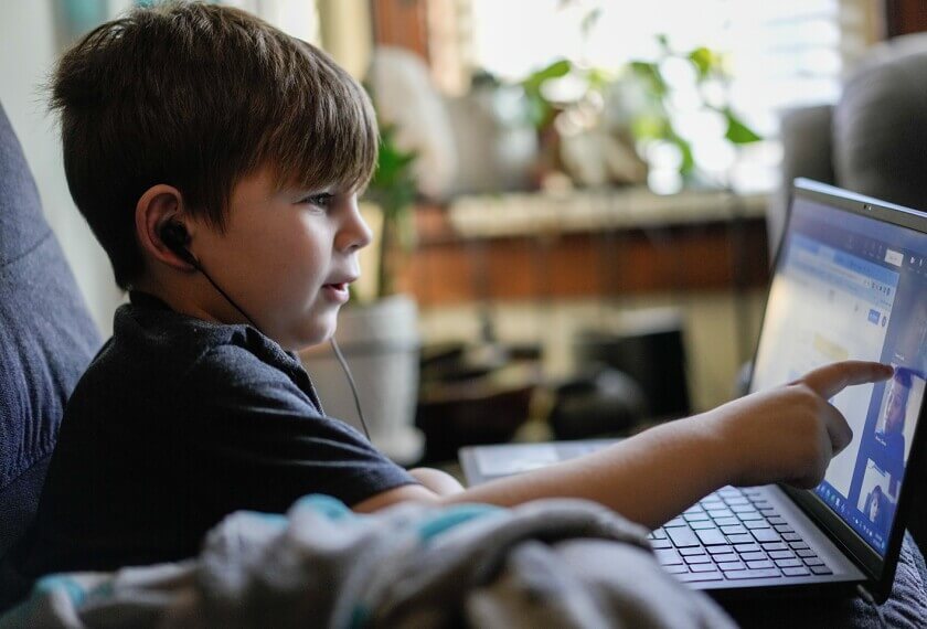 A child sits on a couch, wearing earphones, engaged with a laptop, pointing at the screen, surrounded by indoor plants.