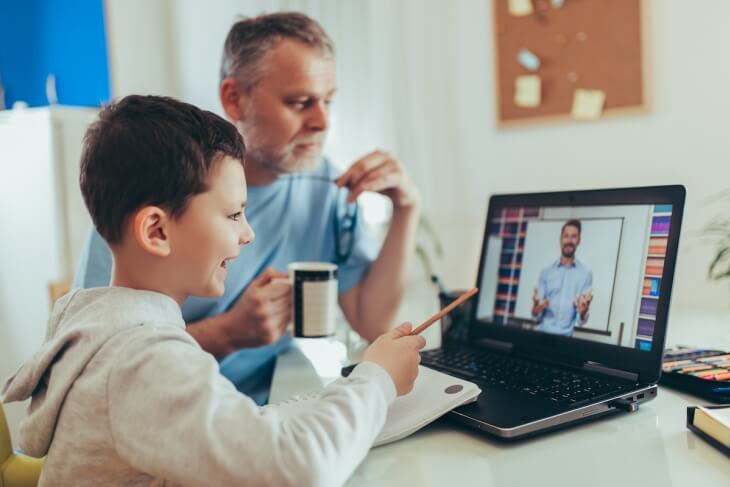 A child and an adult engage in an online learning session, with a laptop displaying a teacher on the screen and notebooks on the table.