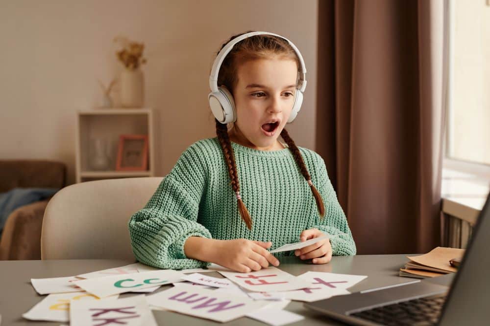 A child wearing headphones sits at a desk surrounded by colorful letter flashcards, focused on learning. A laptop is also on the table.