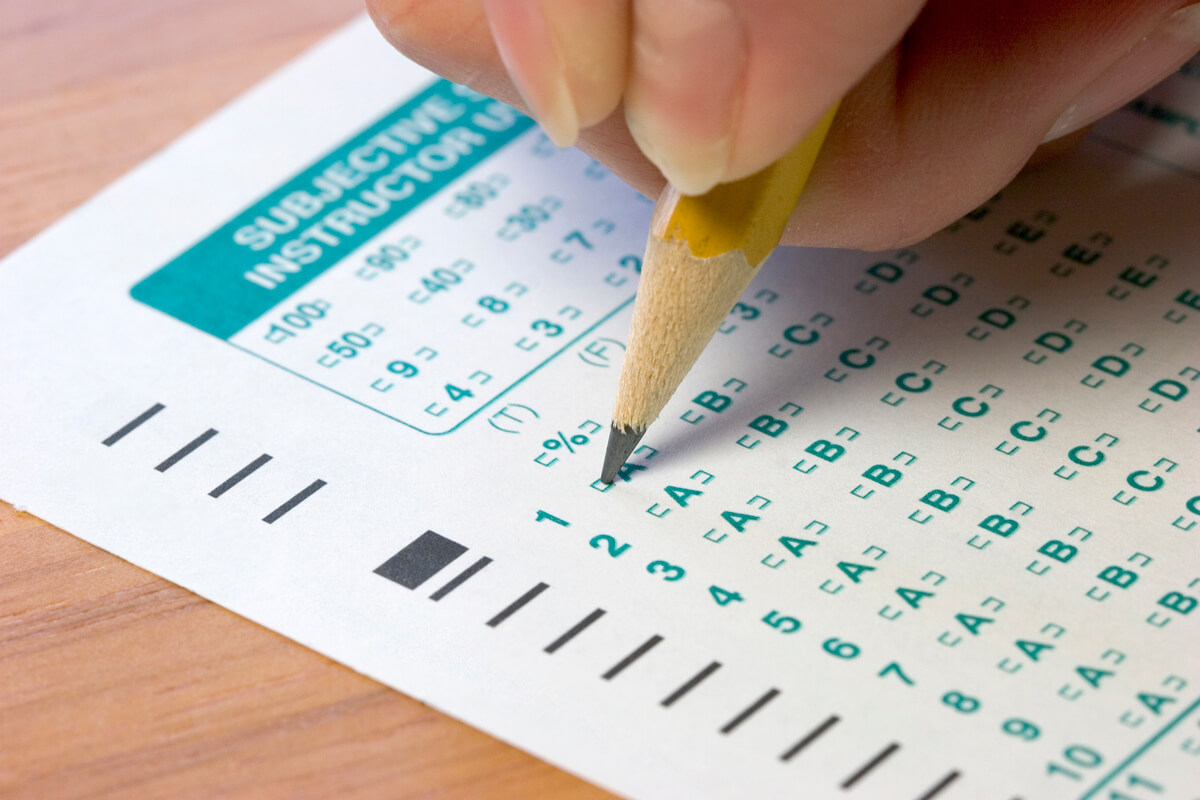 A hand holding a pencil marks answers on a scantron sheet with multiple choice options and a percentage score section.