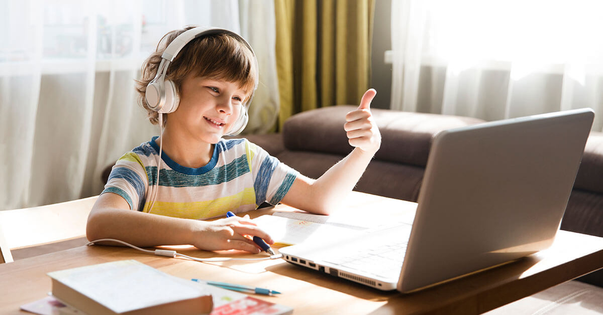 A child wearing headphones gives a thumbs up while sitting at a table with a laptop and school materials, bathed in natural light.