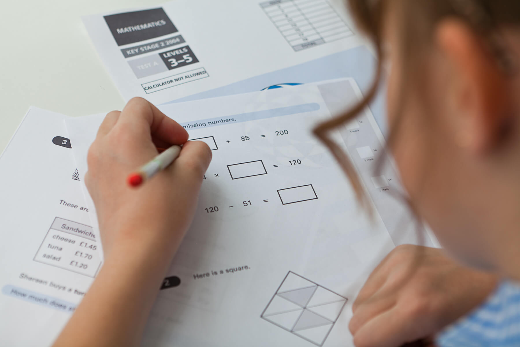 A person's hand holding a pen writes on a math worksheet, filled with equations and geometric shapes. Papers and a calculator are in view.