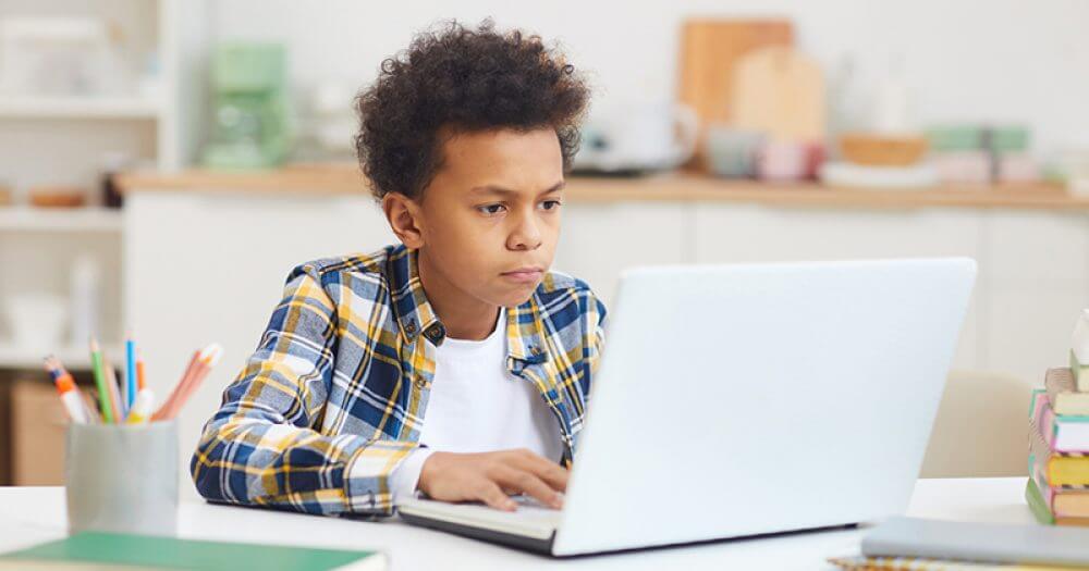 A child in a plaid shirt sits at a desk, engaging with a laptop in a cozy, well-lit kitchen environment.