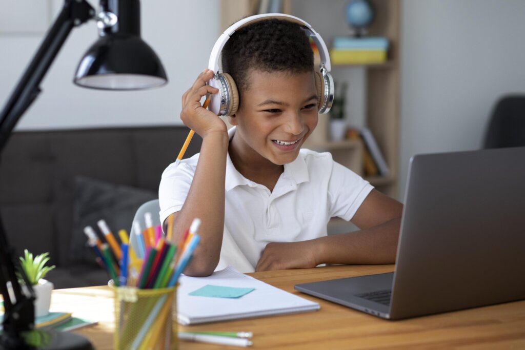 A child wearing headphones sits at a desk, focused on a laptop, with colorful pencils and a notebook in the foreground.