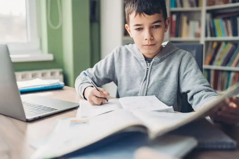 A child in a gray hoodie studies at a wooden desk, surrounded by open textbooks and a laptop, focused on their work.