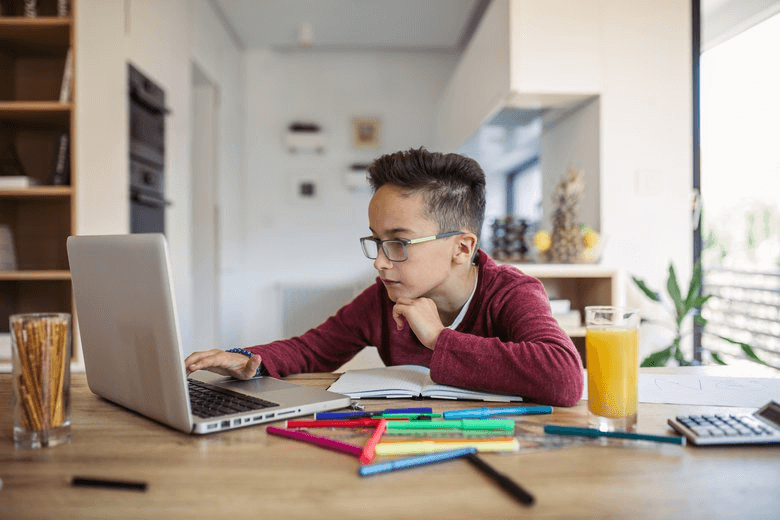 A child sits at a table with a laptop, notebooks, colorful markers, and a glass of orange juice in a bright, airy room.