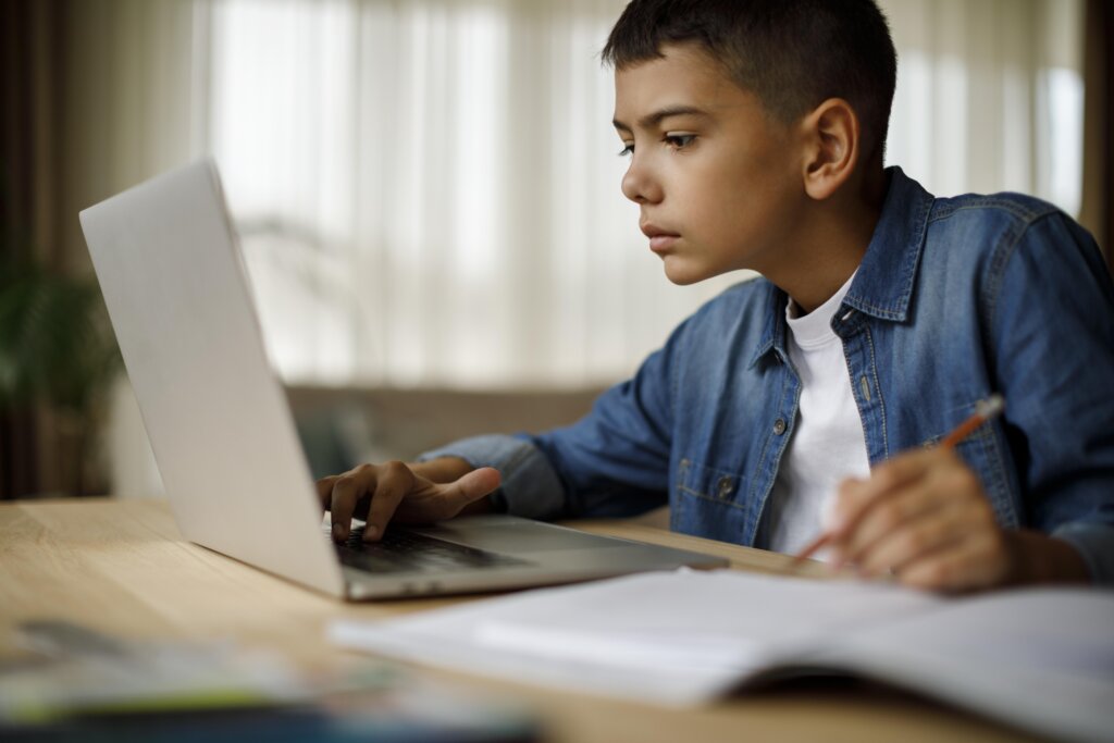 A person in a denim jacket focuses on a laptop while holding a pencil, surrounded by books on a wooden desk. Natural light filters in.