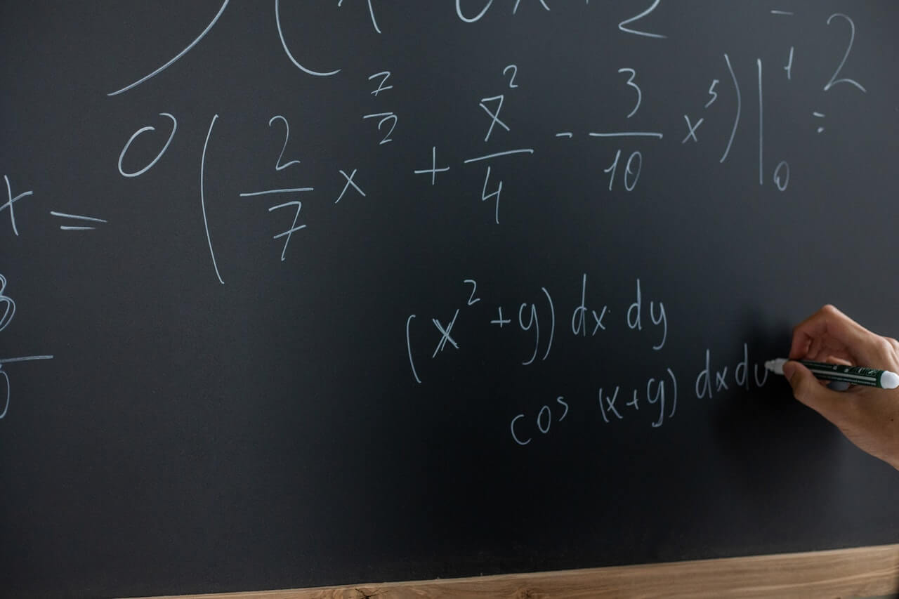 A hand writes mathematical equations in white chalk on a blackboard, with various integrals and functions displayed prominently.
