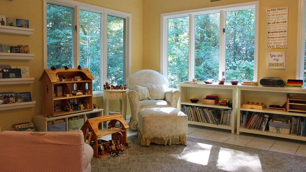 Cozy reading nook with a plush chair, toy shelves, and large windows overlooking a lush green forest. Ideal for relaxation and play.