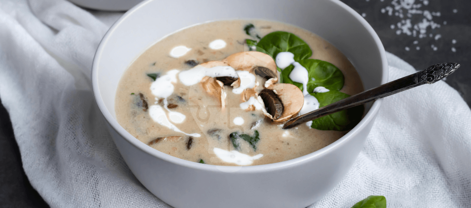 Creamy mushroom soup garnished with sliced mushrooms and spinach leaves in a white bowl, on a white cloth, exuding a cozy, hearty feel.