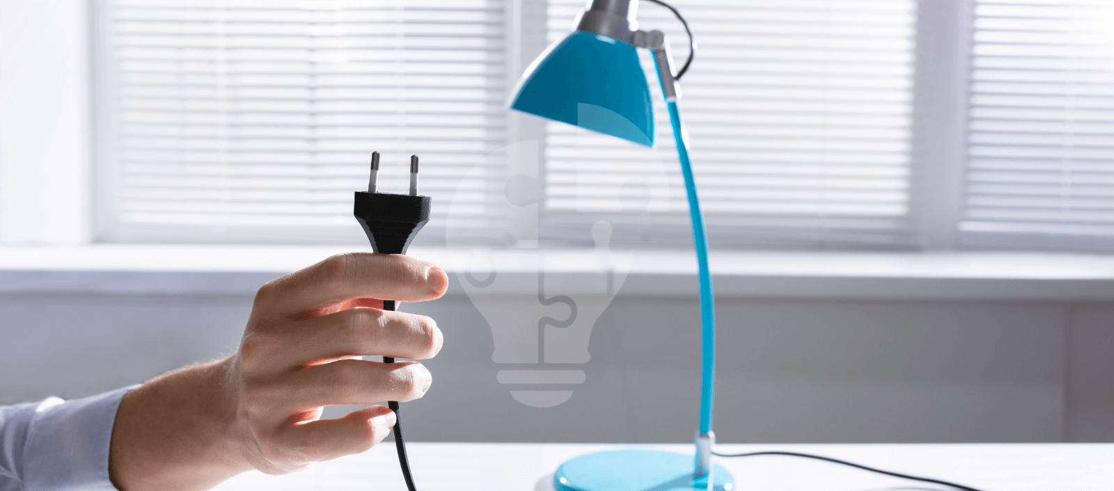 A hand holding a black electrical plug near a blue desk lamp, with light filtering through blinds in the background.