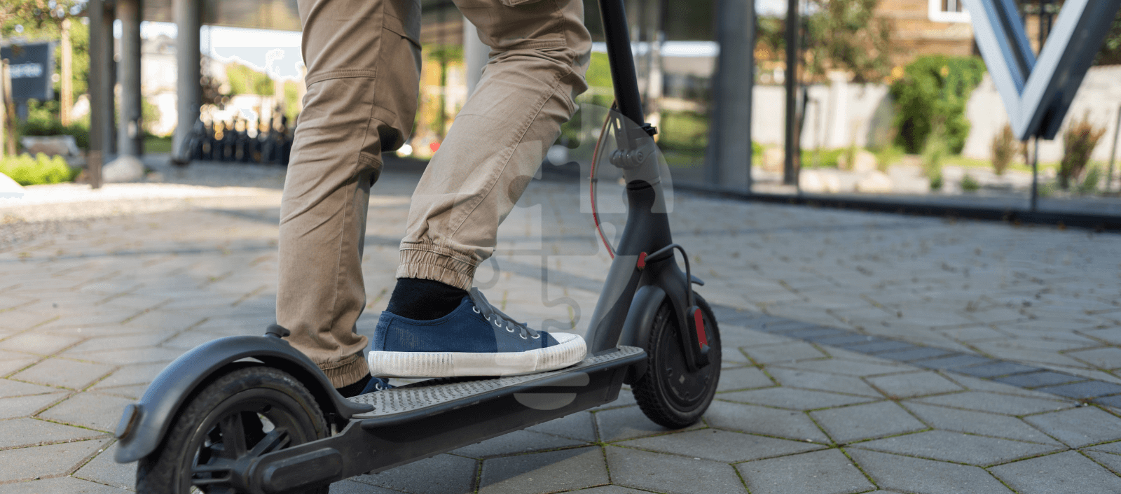 A person wearing beige pants and blue sneakers rides a black electric scooter on a tile path. The scene has a modern, urban feel with glass buildings in the background.