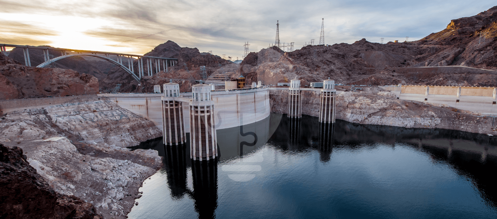 Hoover Dam at sunset with reflected towers, surrounded by rocky terrain and the Colorado River, under a cloudy sky.